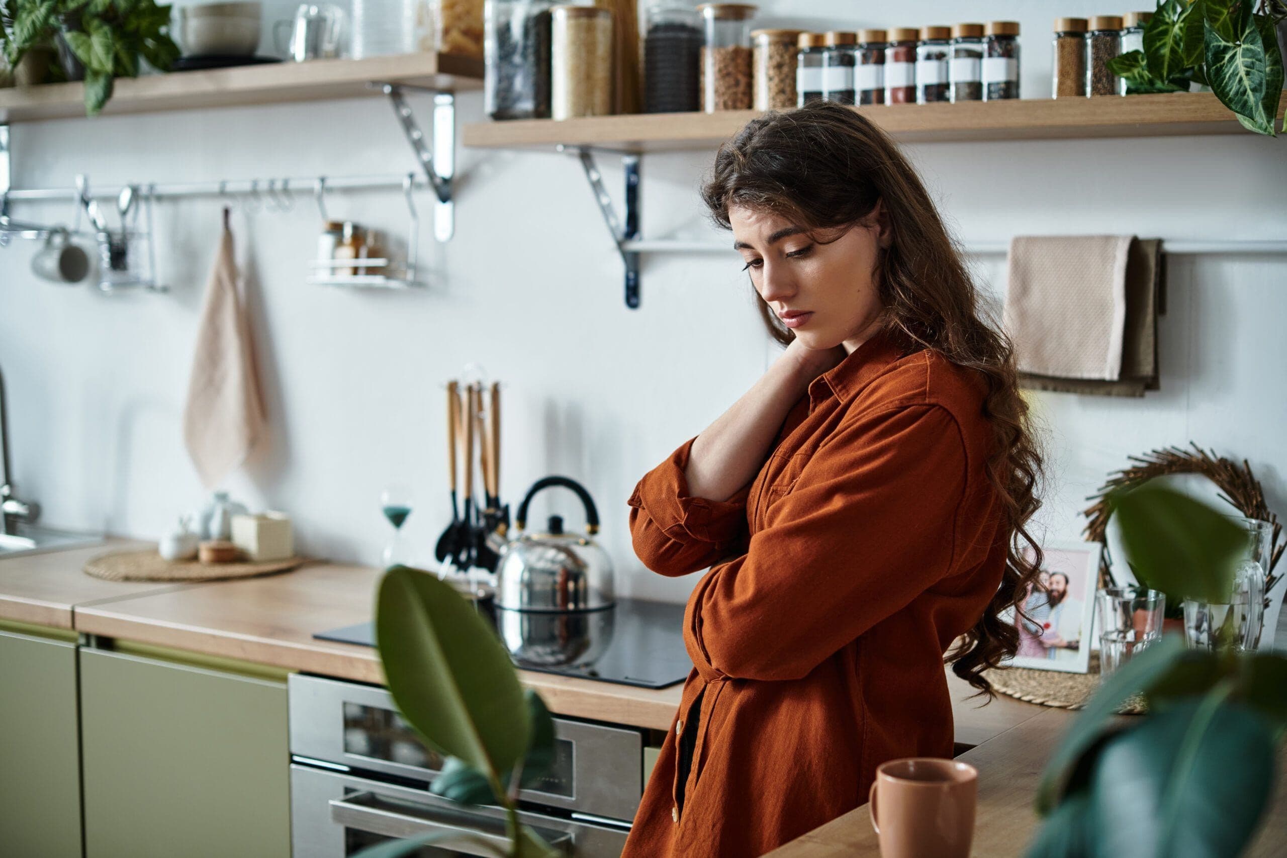A woman stands in her kitchen, hand on her neck, lost in thought and sadness.