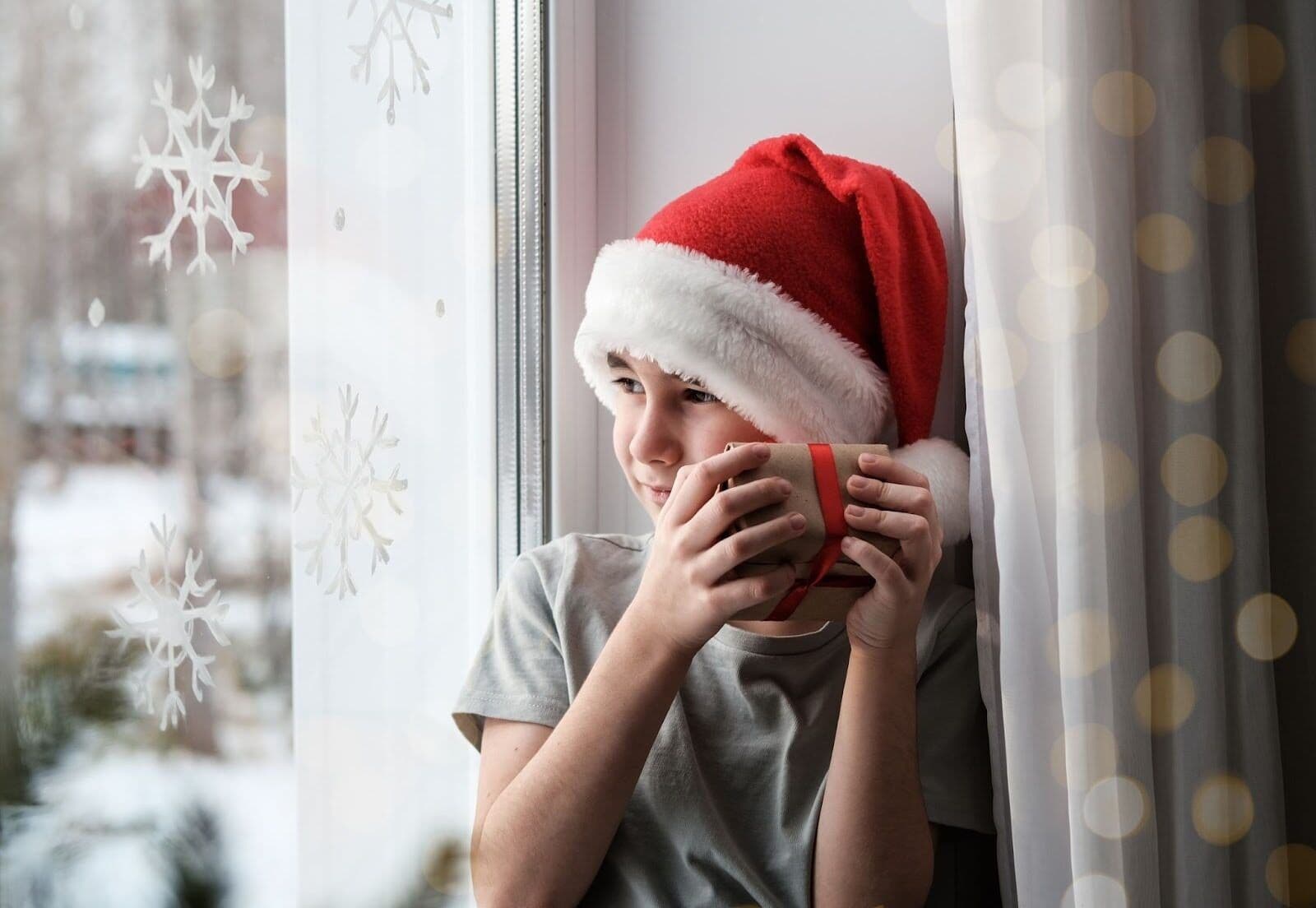 A child wearing a Santa hat holding a gift while looking out the window, symbolizing grief at Christmas and the emotional weight children may feel during the holidays.