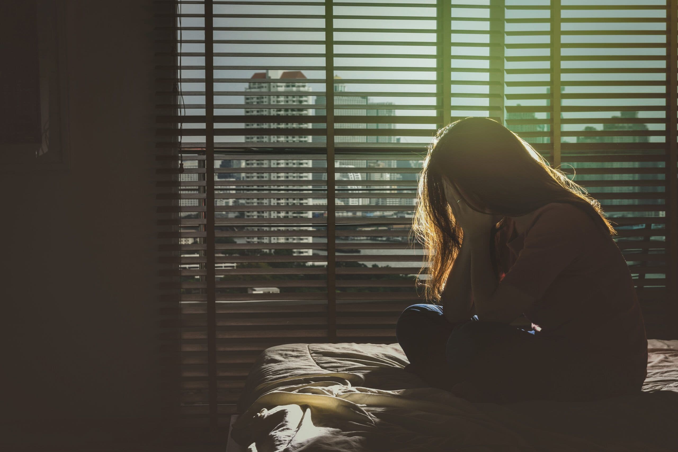 A person sits alone in a dimly lit room, head in hands, reflecting the grief and isolation felt by international survivors of suicide loss.